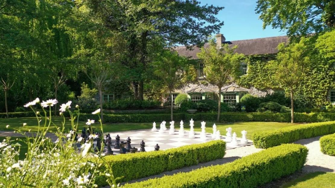 Outdoor chessboard with large black and white chess pieces in a garden with a stone building covered in ivy.