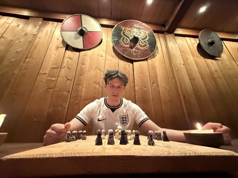 Ed Southgate studying Viking chess, sitting at a wooden table with a lit candle, wearing a white England jersey, surrounded by wooden walls with Viking shields.