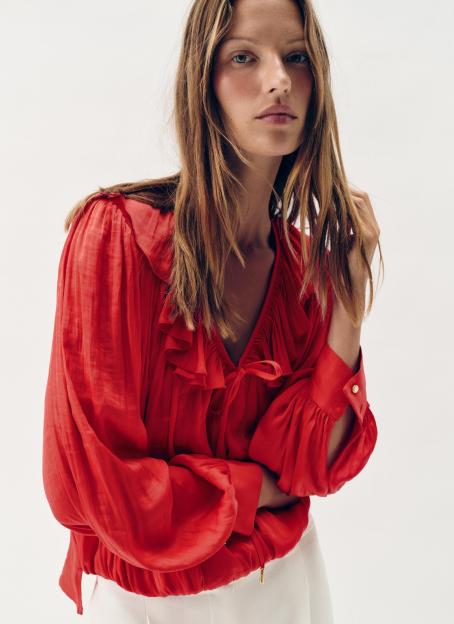 A model wearing a red long-sleeved blouse with ruffles and a tie-neck, paired with white pants.