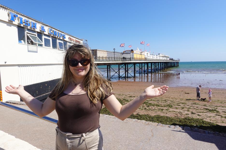 A woman with sunglasses stands in front of a white building with "Fish & Chips" written on it and a pier extending into the sea on a sunny day.