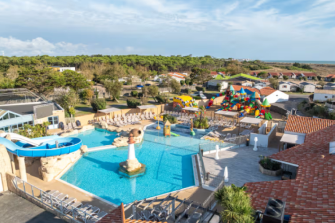 Aerial view of the Sol a Gogo campsite in St Hilaire-de-Riez, France, showing a swimming pool with a lighthouse and waterslide, a children's playground, and mobile homes, with a pine forest and the ocean in the background.