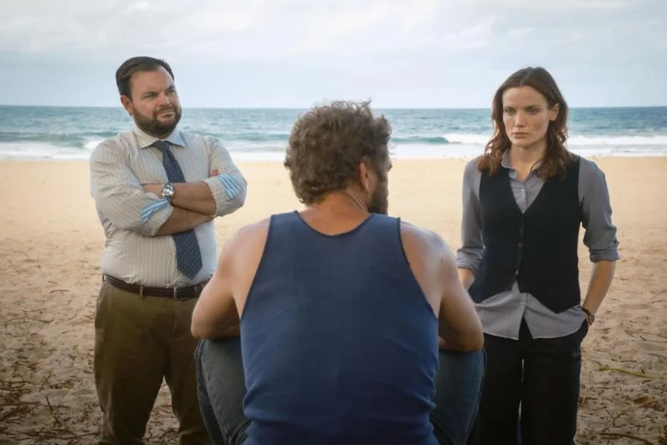 Detective Colin Cartwright (Lloyd Griffith) and DI Mackenzie Clarke (Anna Samson) stand on a beach with the ocean in the background, looking down at a man seen from behind.