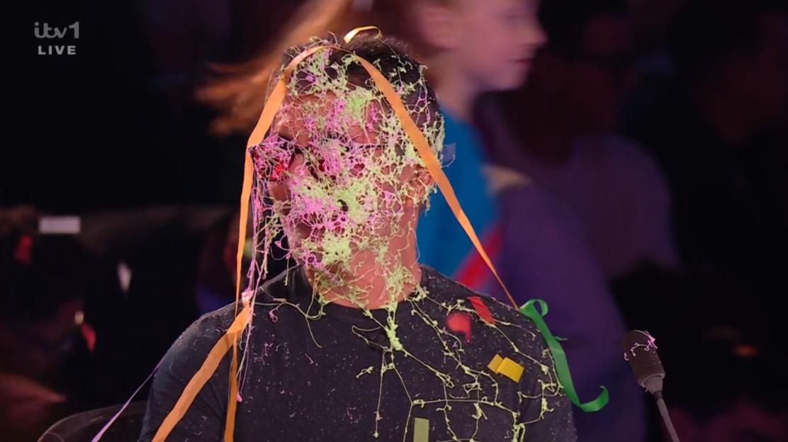 Simon Cowell's face and shirt covered in silly string and streamers.