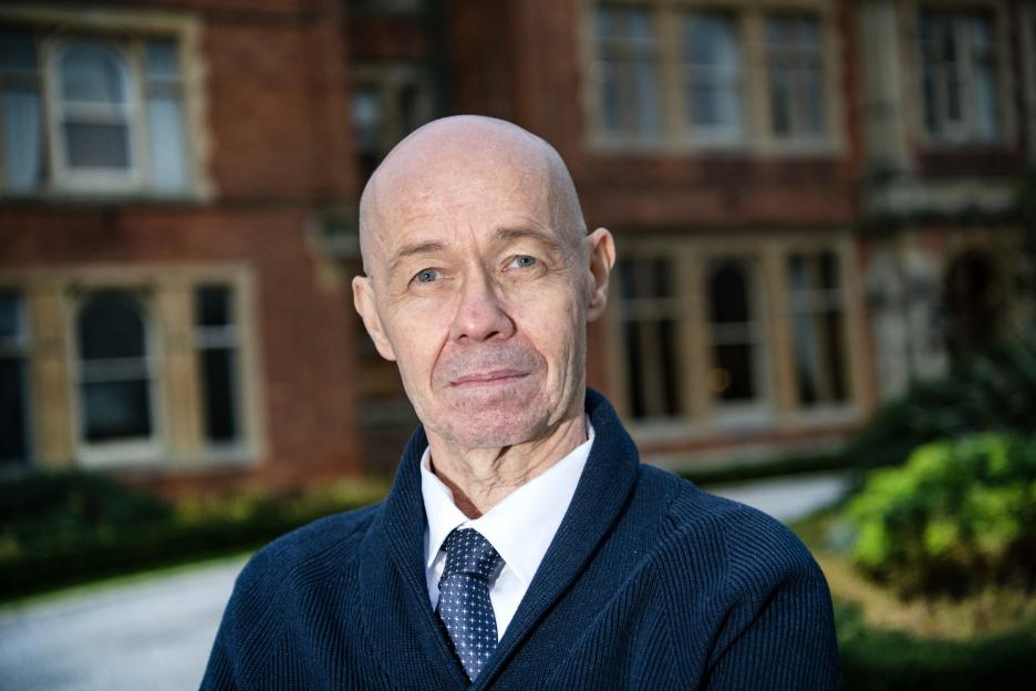 Albert Douglas, a bald man with light eyes, wearing a dark blue cardigan and a patterned blue tie over a white shirt, stands outdoors with a brick building in the blurred background.