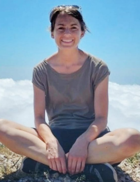 Lisa Ross, a woman in a grey t-shirt and sunglasses on her head, smiles at the camera while sitting cross-legged outdoors with a cloudy sky behind her.