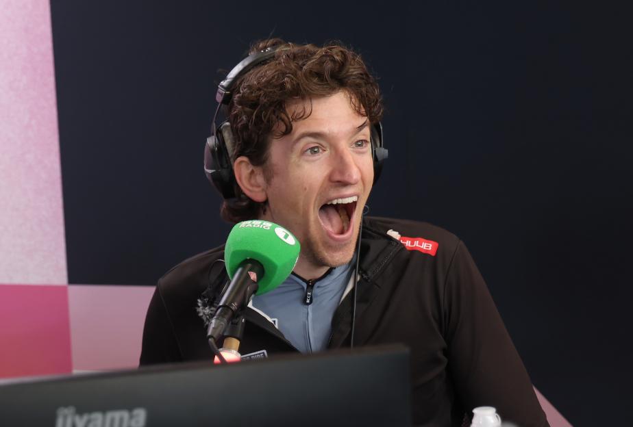 Greg James reacting in a Radio 1 interview, wearing headphones and speaking into a green microphone.