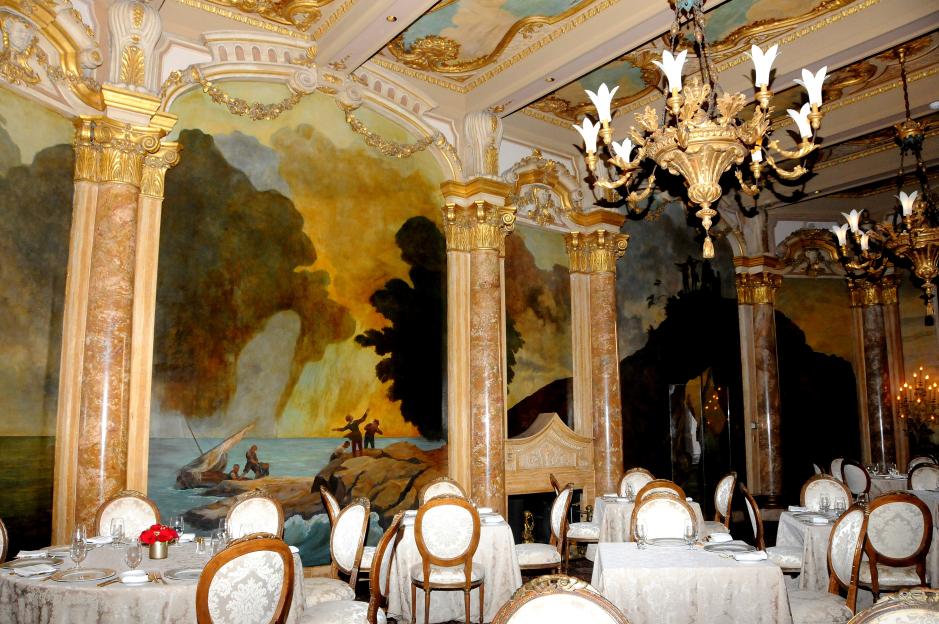 Interior view of the dining room at the Mar-a-Lago estate, with ornate gold decor, large chandeliers, set dining tables, and murals on the walls.