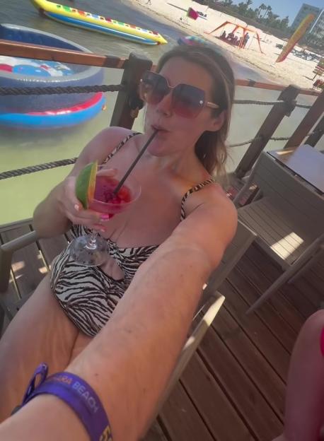 Becki Jones in a zebra-print bikini, drinking a cocktail on a pier overlooking a beach.