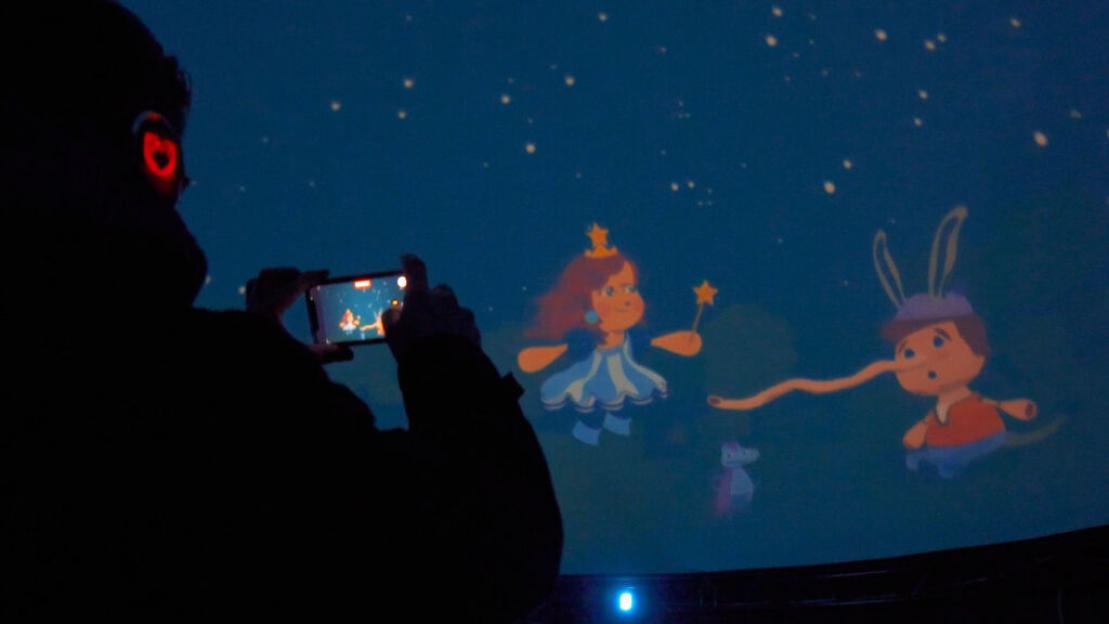 A person in a planetarium uses their phone to record an animated scene of a fairy and Pinocchio projected onto the dome.