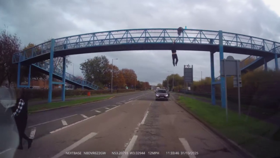 A person dangling from a blue pedestrian bridge over a road, with their hands gripping the edge and legs hanging freely in the air.