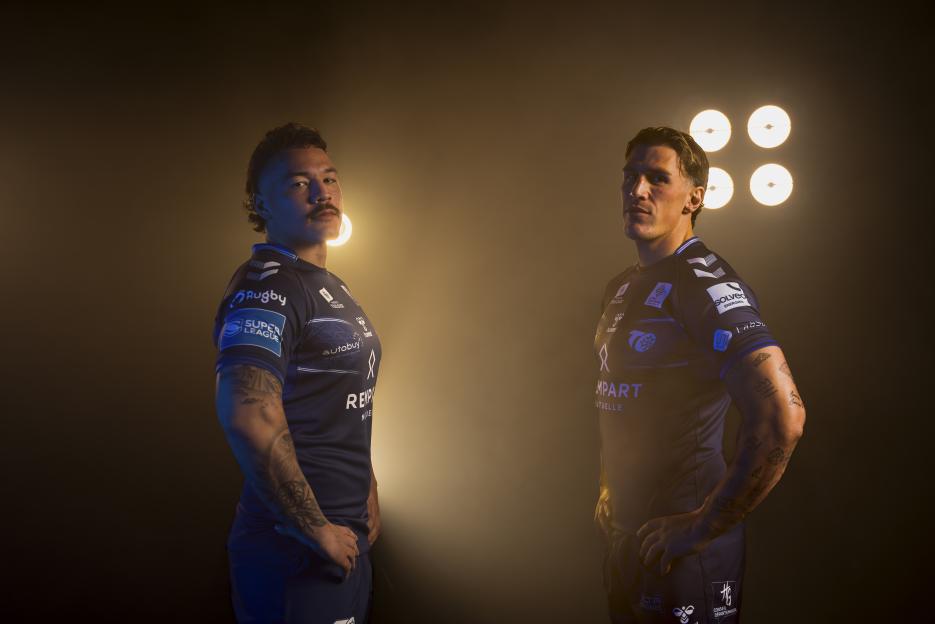 Two rugby players in uniform, Tyler Dupree (left) and another player, posing with hands on hips against a dark background with bright lights.