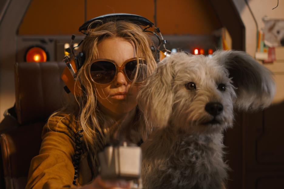 Milly Alcock wearing headphones and sunglasses, holding a device, with a fluffy white dog beside her, in a scene from "Supergirl."