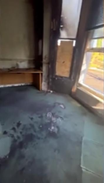 Damage to a building from a petrol bomb attack, with a broken window boarded up and charring on the frame.