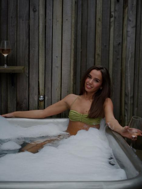 A woman in a green bikini top smiles while relaxing in a hot tub filled with bubbles, holding a glass of wine.