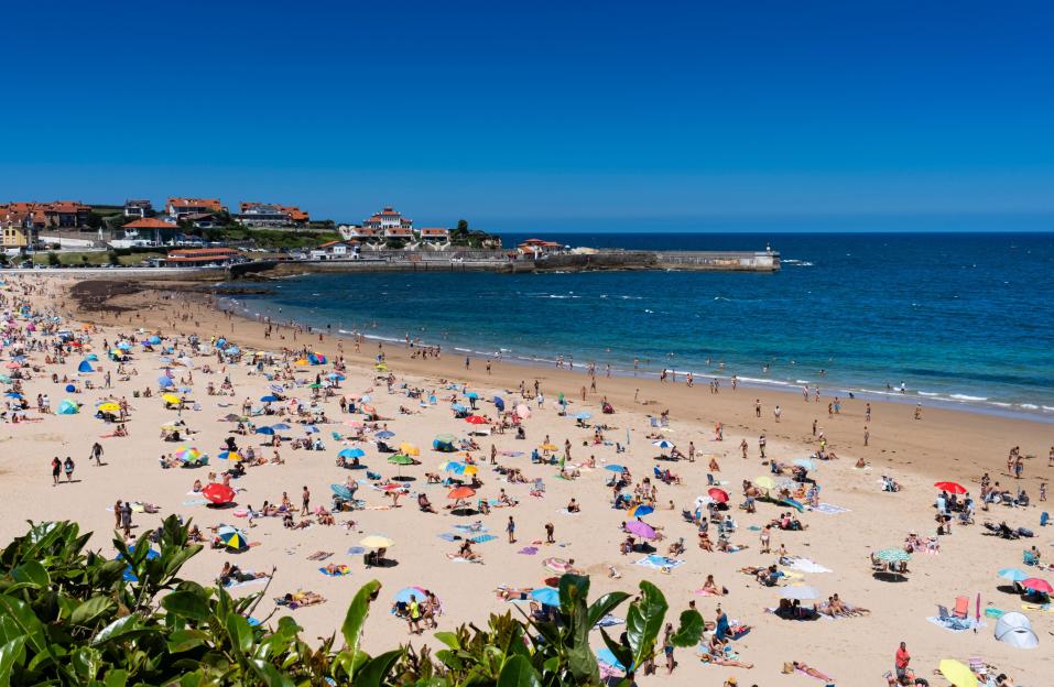 Aerial view of Comillas beach on a sunny summer day with blue sky, showing many people sunbathing under colorful umbrellas.