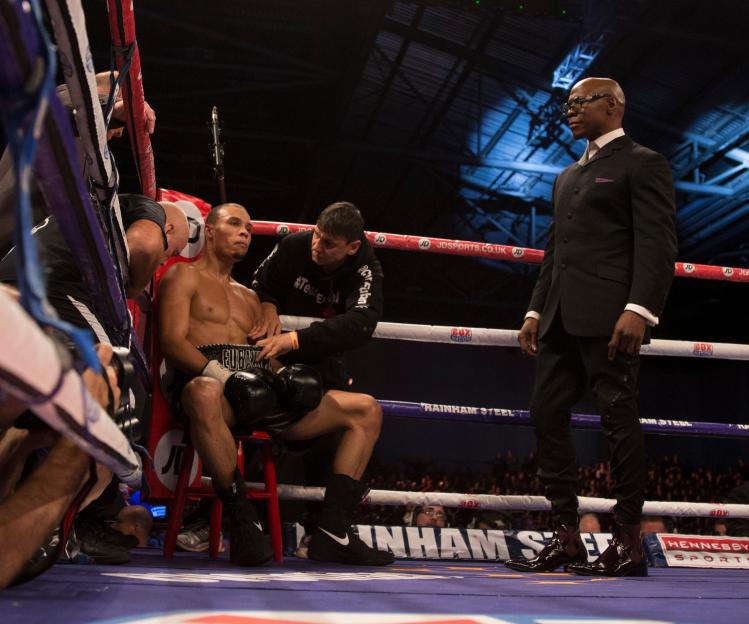 Boxer Chris Eubank Jr. sits in the corner of a boxing ring while his trainer tends to him, with Chris Eubank Sr. standing nearby.
