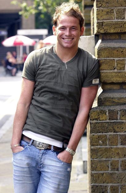 Actor Joe Swash smiling while leaning against a brick wall.