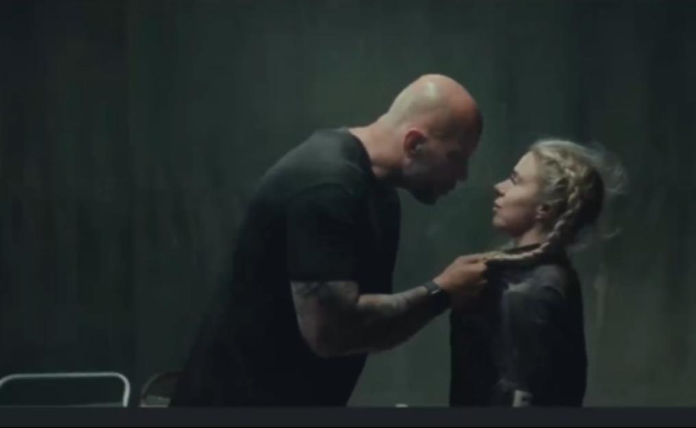 A bald man in a black shirt holding a blonde woman by her braided hair.