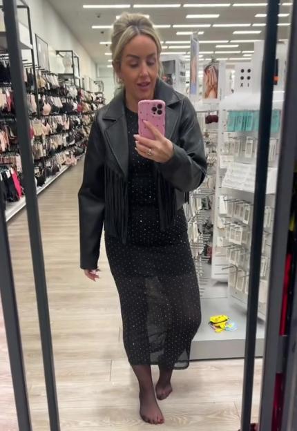 A woman wearing a black dress, fringed leather jacket, and pantyhose, holding a pink phone, reflected in a mirror inside a store.