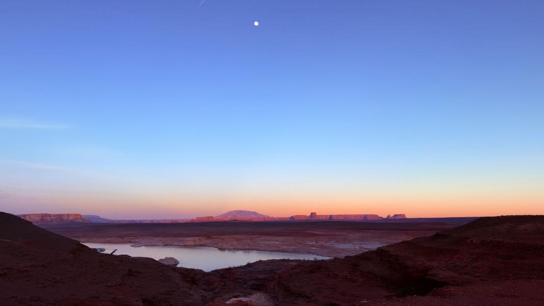 Panoramic view of a desert landscape at dusk with a pale full moon in a blue sky.