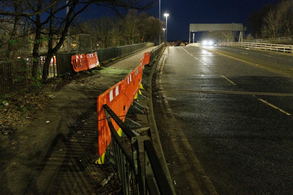 A street scene at night with orange safety barriers along the roadside and headlights in the distance.
