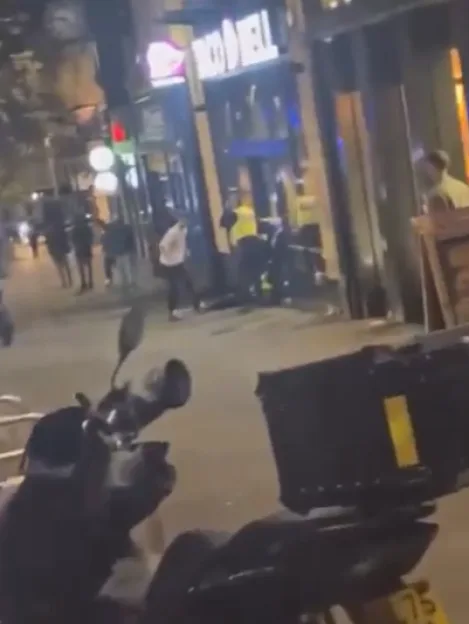 A low-quality, blurry, street-level video still showing a nighttime scene outside a storefront, with a person lying on the ground in the distance and a delivery motorcycle parked in the foreground.
