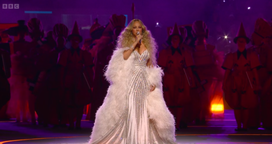 Mariah Carey performing on stage in a glittering gown and a feather boa, with backup performers in conical hats and red outfits behind her.
