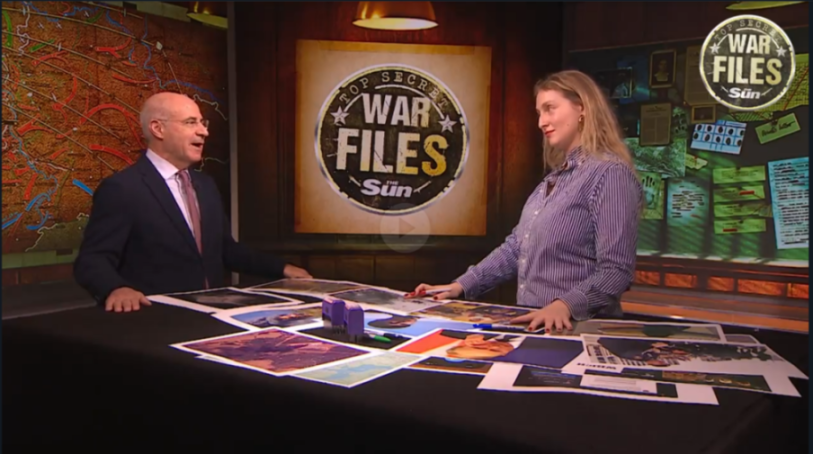 A man and a woman stand behind a table covered with photos and papers, facing each other in a studio set with a "Top Secret War Files" logo in the background.
