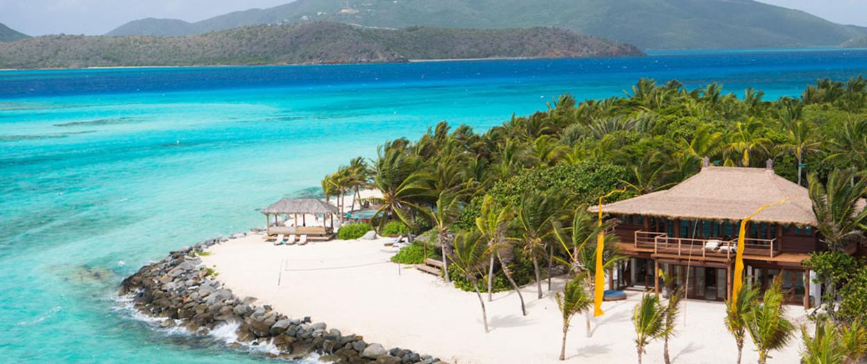 A large wooden house with a thatched roof, surrounded by palm trees on a white sand beach next to turquoise water.