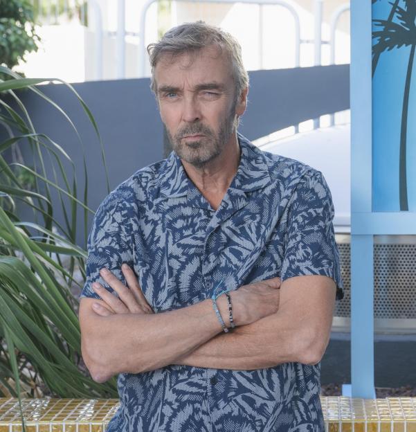 John Hannah as Dennis from Benidorm Is Murder, standing with arms crossed next to a sign for "Tonight Over 50's Singles Night".