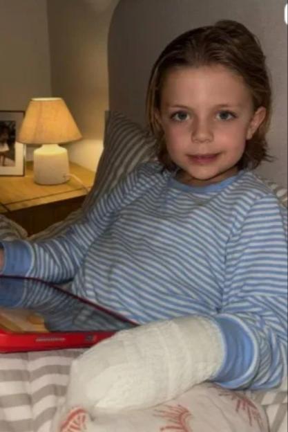 A young child with a bandaged arm, wearing pajamas and holding a red tablet in bed.