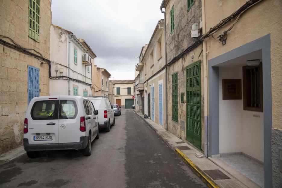 A quiet street in the village of Santa Margalida, Majorca, with several vans parked on the left side and tall buildings with green and blue shutters lining the narrow road under a cloudy sky.