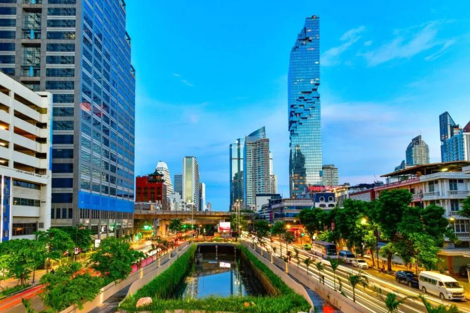 Night view of the Bangkok metropolitan skyline, featuring modern skyscrapers, commercial buildings, and illuminated streets alongside a canal at dusk.