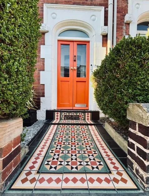 A pathway with patterned tiles leading to a bright orange double front door of a brick house.