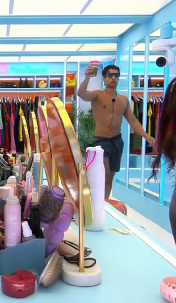 A man with dark sunglasses and a bare chest stands in a dressing room, holding up a drink.