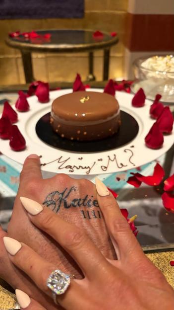 Close-up of a hand with a "Katie" tattoo on its top, pointing to a cake that says "Marry Me?" The hand is wearing a large engagement ring, and is surrounded by rose petals.