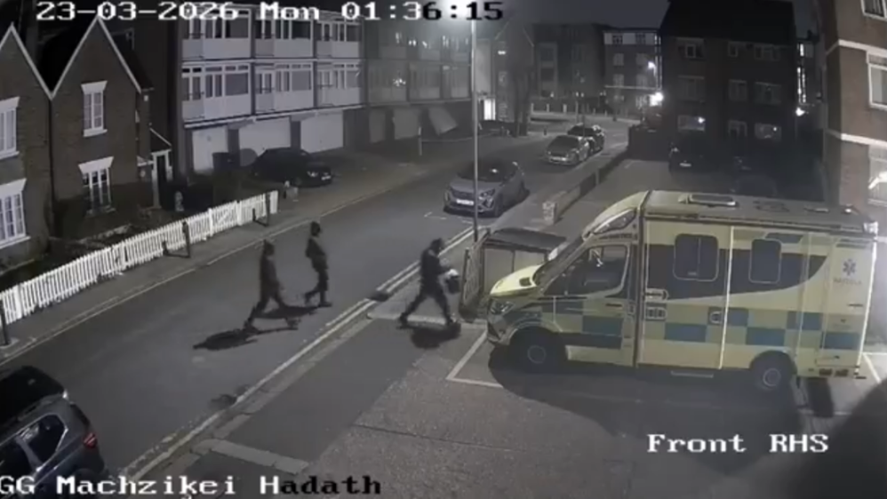 Three people walk past an ambulance parked on a residential street at night.