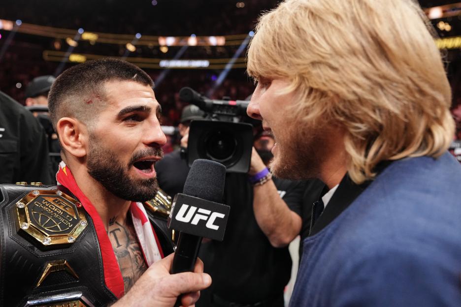 Ilia Topuria wearing a championship belt and speaking into a UFC microphone while facing Paddy Pimblett.