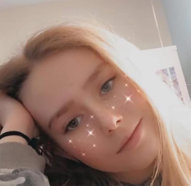 A young girl with blonde hair, wearing a camouflage hoodie, with sparkle filter on her face.