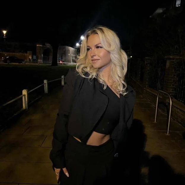 A blonde woman in a black cropped top and jacket stands on a street at night.