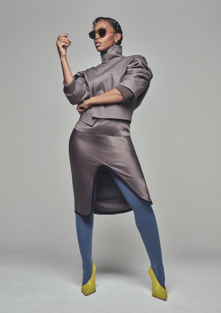 Nafessa Williams in a gray high-neck top, gray skirt, blue tights, and yellow heels.
