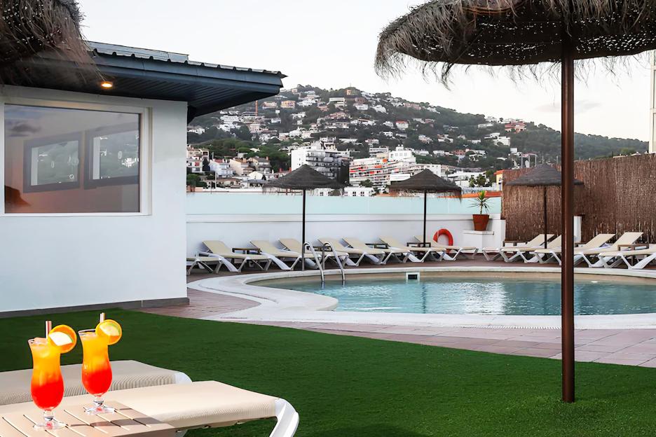 A hotel rooftop pool with lounge chairs, two cocktails, and a view of a town built into a hill.