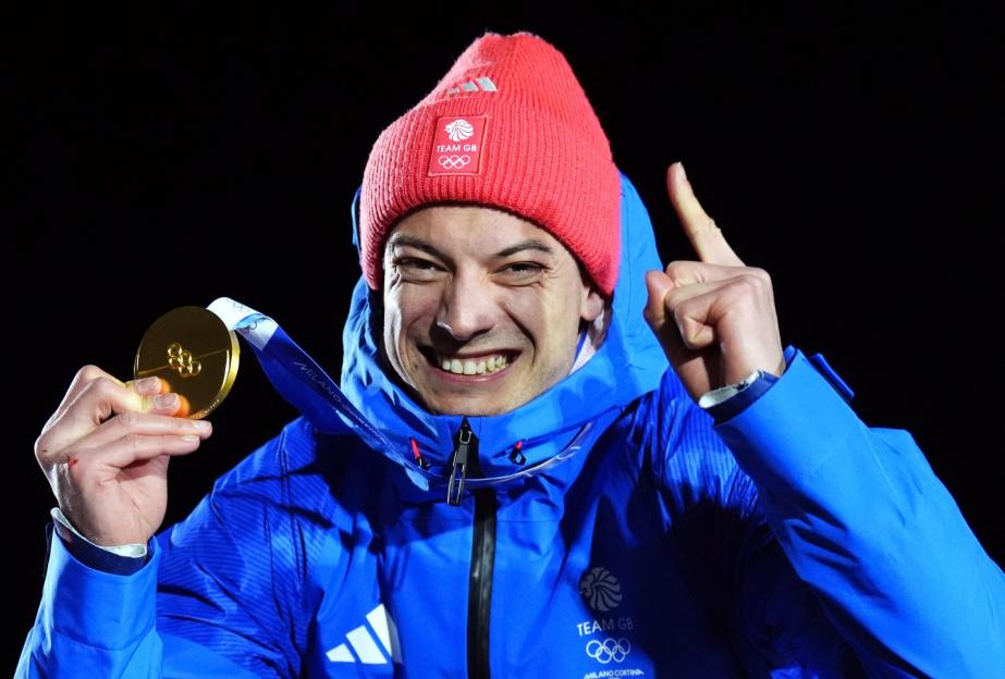 A smiling athlete in a blue jacket and red beanie, holding a gold medal and pointing a finger up.