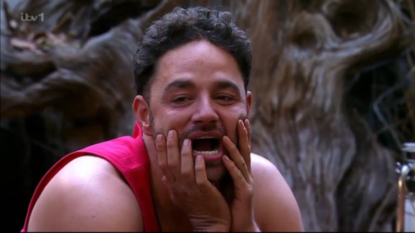 Man in a pink vest with hands covering his cheeks and mouth open in a surprised expression.