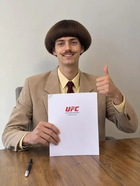 A smiling man with a bowl cut and mustache wearing a suit holds a UFC contract and gives a thumbs-up.