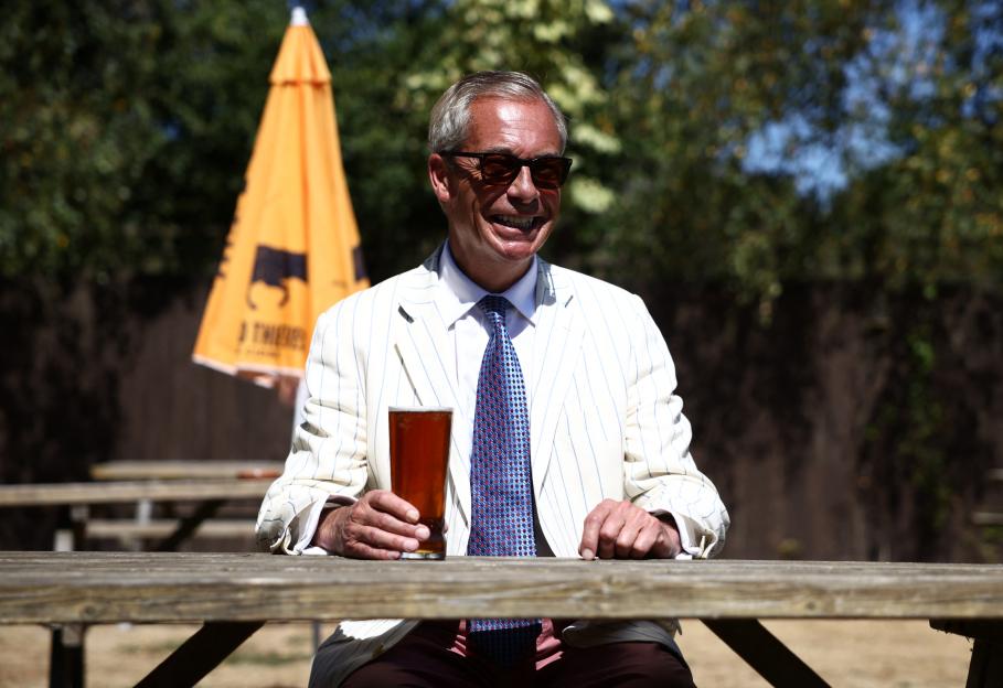 Nigel Farage, leader of Reform UK, sits at an outdoor picnic table with a pint of beer in hand, smiling.