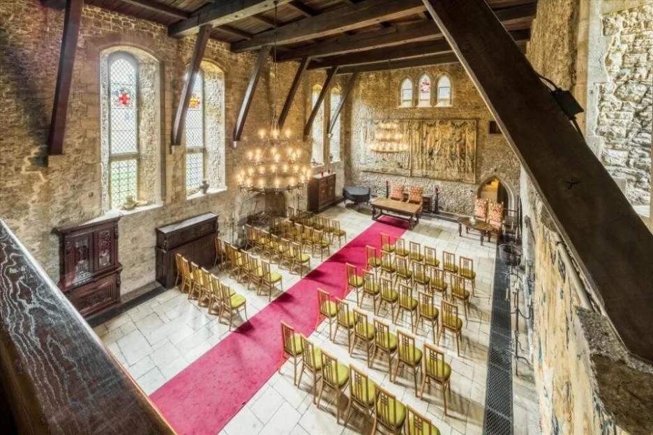 High-angle view of a medieval great hall with stone walls, wooden beams, a red carpet down the aisle, and rows of chairs set up for an event.