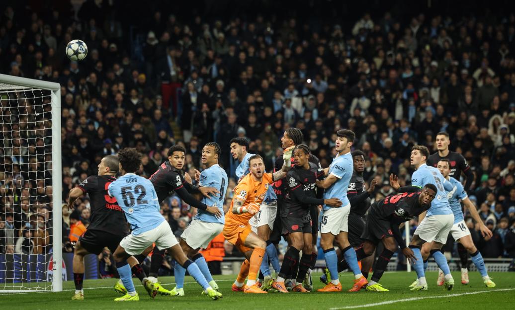 A soccer ball hovers above the goal as players from Manchester City and Bayer Leverkusen vie for position.