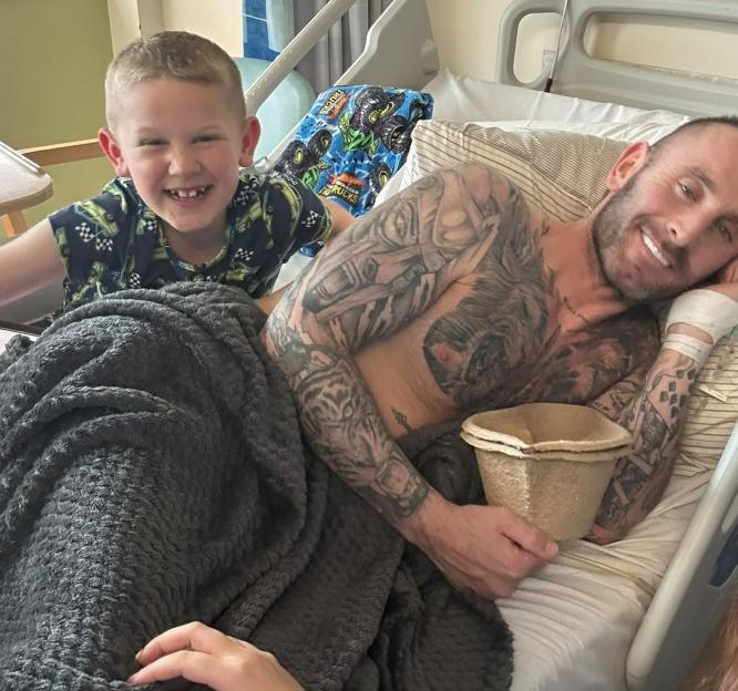 James Bainbridge, a man with numerous tattoos, smiles from a hospital bed next to his young son, Alfie, who also smiles.