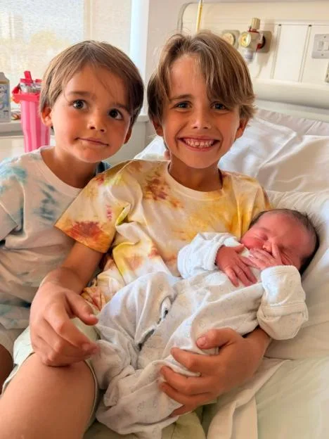 Two young boys smiling while holding a newborn baby in a hospital bed.